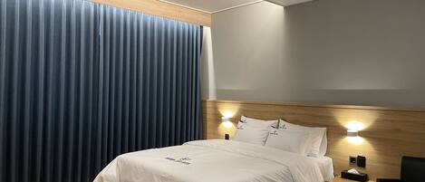 Una habitación de hotel moderna con una cama grande, cabecera de madera, dos lámparas de noche, una mesita de noche, un escritorio y un ventanal con cortinas azules.