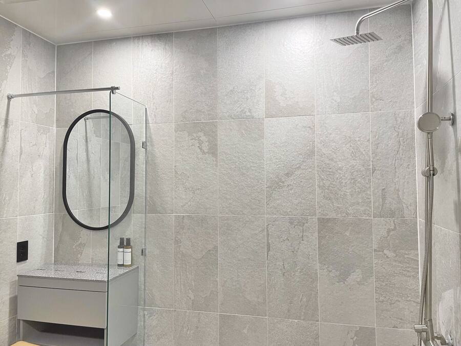 Un baño moderno con azulejos grises, una ducha con mampara de vidrio y un mueble de tocador con espejo y artículos de aseo.
