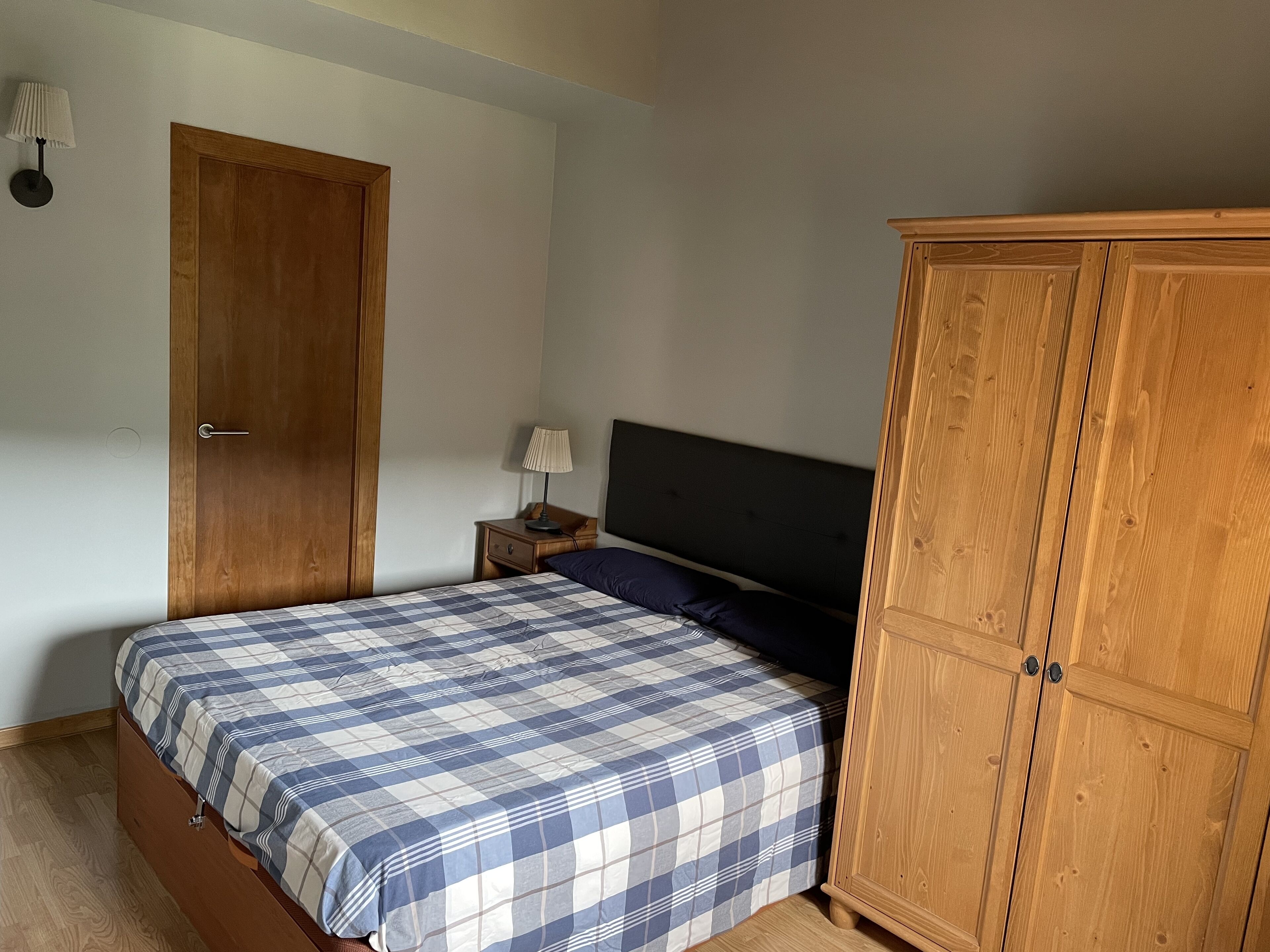 A bedroom with a bed, a wooden wardrobe, a nightstand, and a wall-mounted lamp.