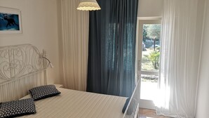 A bedroom with a bed, a hanging light fixture, a metal headboard, and a view of the outdoors through a window with curtains. - Apartment Dory (Malinska-Dubasnica)