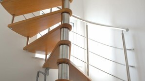 A spiral staircase with wooden steps and metal railings in a bright room with a white wall and a window. - Apartment Dubravka (Malinska-Dubasnica)
