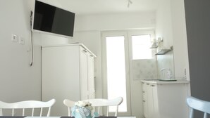 A small, bright room with a white table, two white chairs, a TV mounted on the wall, a door leading outside, and a kitchen area with a sink and cabinets. - Apartment Dubravka (Malinska-Dubasnica)