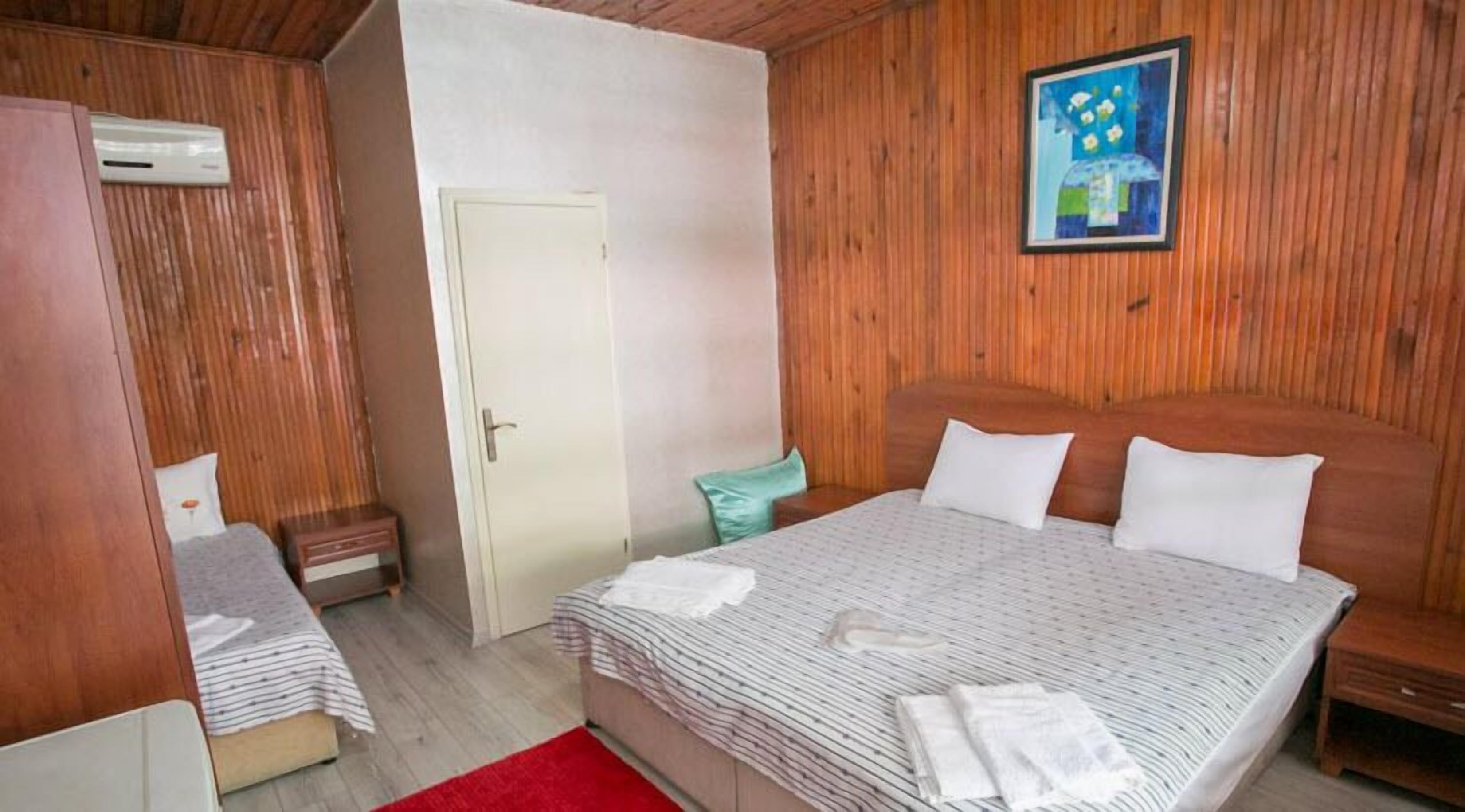 Un dormitorio con cabecera de madera, cama con ropa de cama blanca, alfombra roja, mesita de noche pequeña y aire acondicionado instalado en la pared.