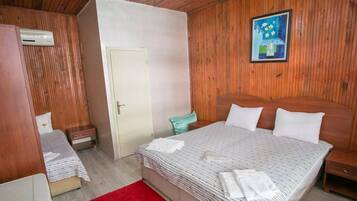 Un dormitorio con cabecera de madera, cama con ropa de cama blanca, alfombra roja, mesita de noche pequeña y aire acondicionado instalado en la pared.