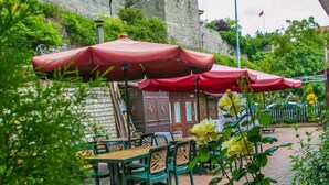 An outdoor café with tables and chairs under red umbrellas, set against a stone wall and greenery. - Carsi Pansiyon (Amasra)