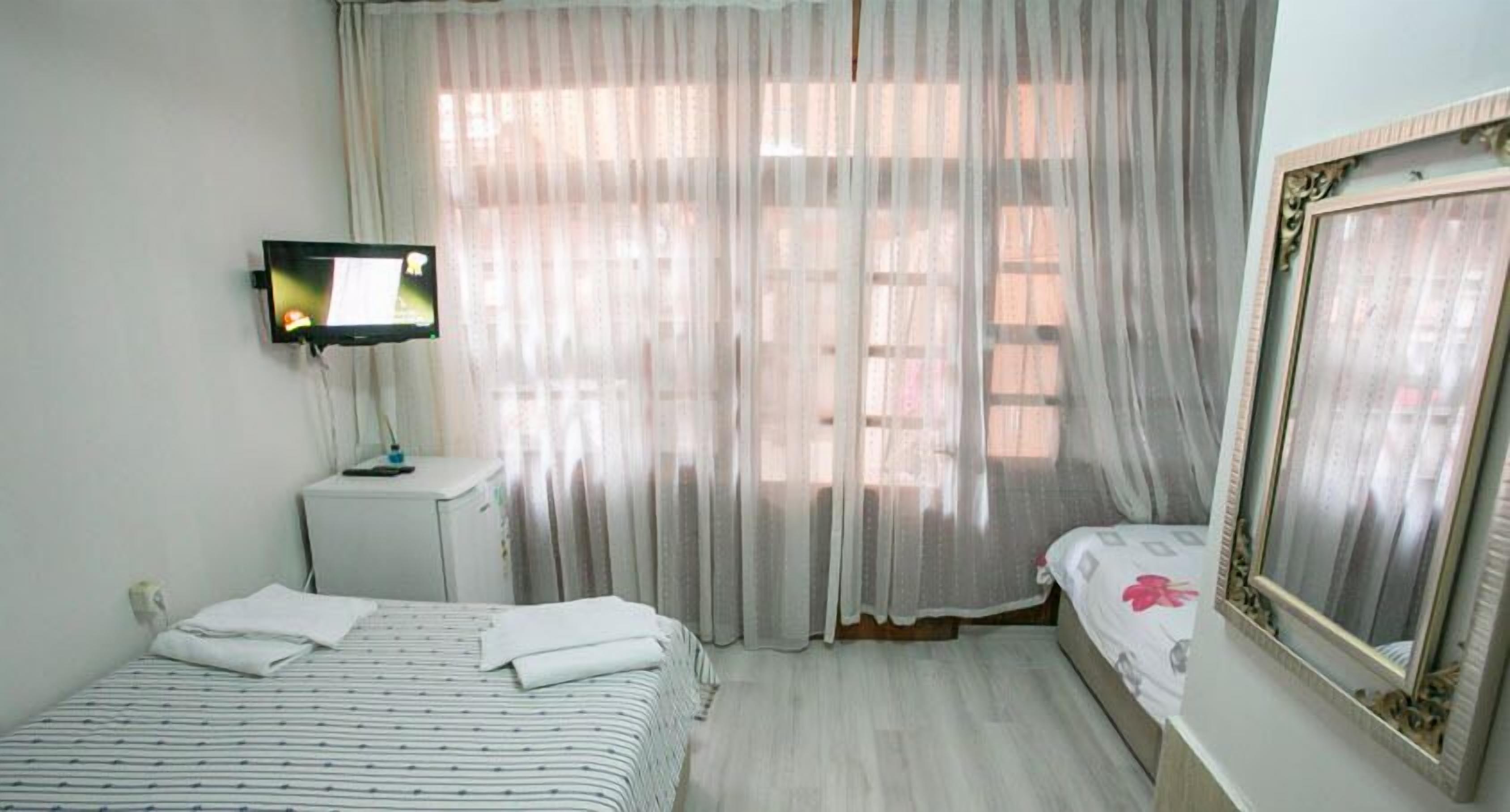 A hotel room with a bed, a bedside table with a lamp, a large window with sheer curtains, and a framed mirror on the wall.