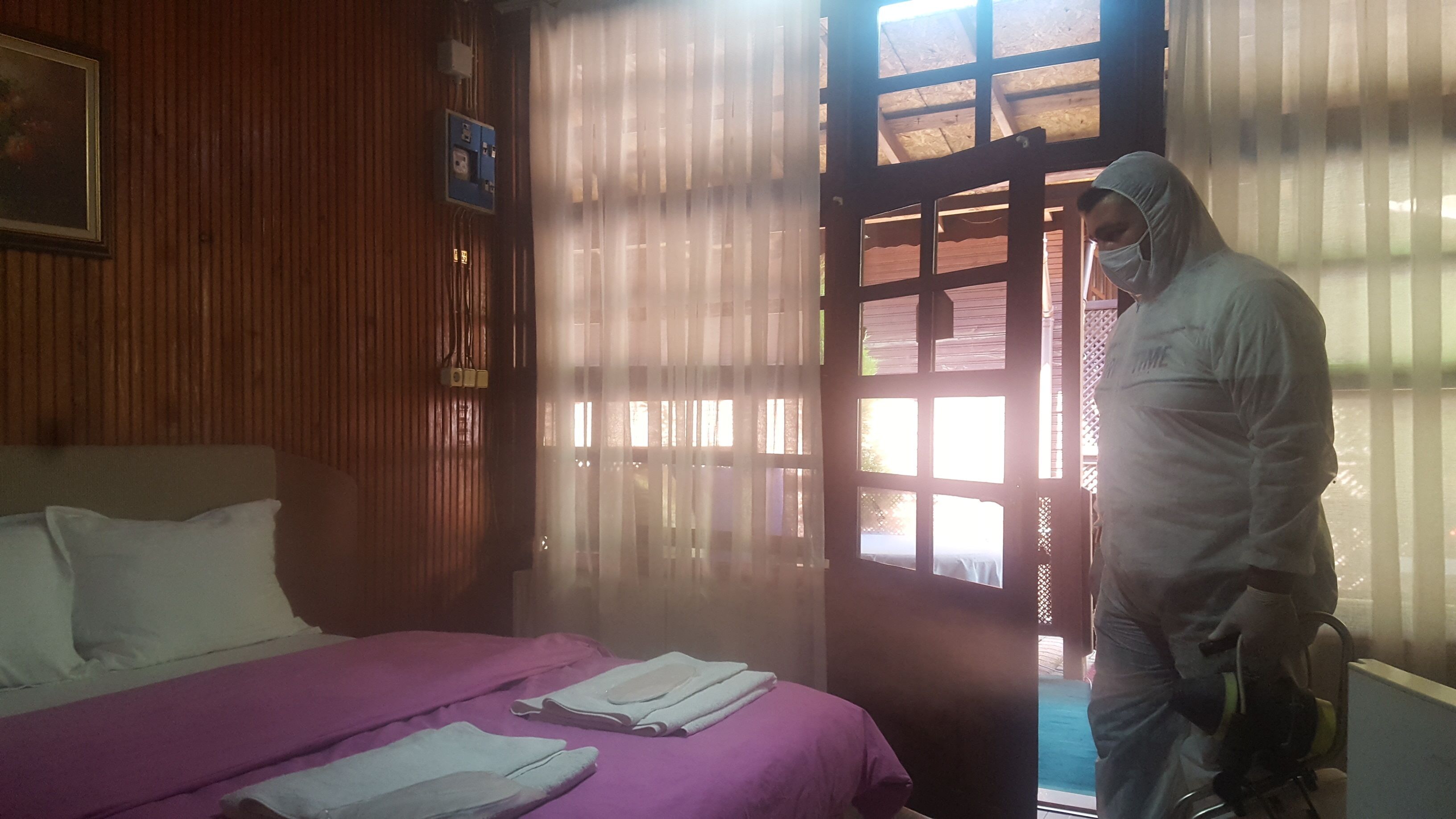 Ein Zimmer mit Bett, einem Fenster mit Vorhängen und einer Person in Schutzkleidung.