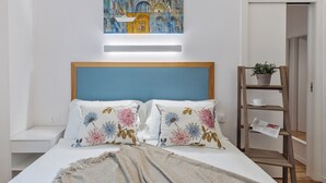 Un dormitorio ordenado con cama, almohadas, un cuadro, una mesita de noche y una estantería de madera con una planta.