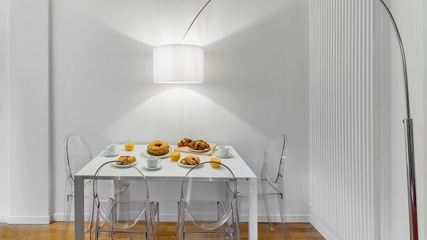 Una mesa de comedor blanca con sillas transparentes, una lámpara blanca y una disposición para el desayuno con pasteles y jugo de naranja.