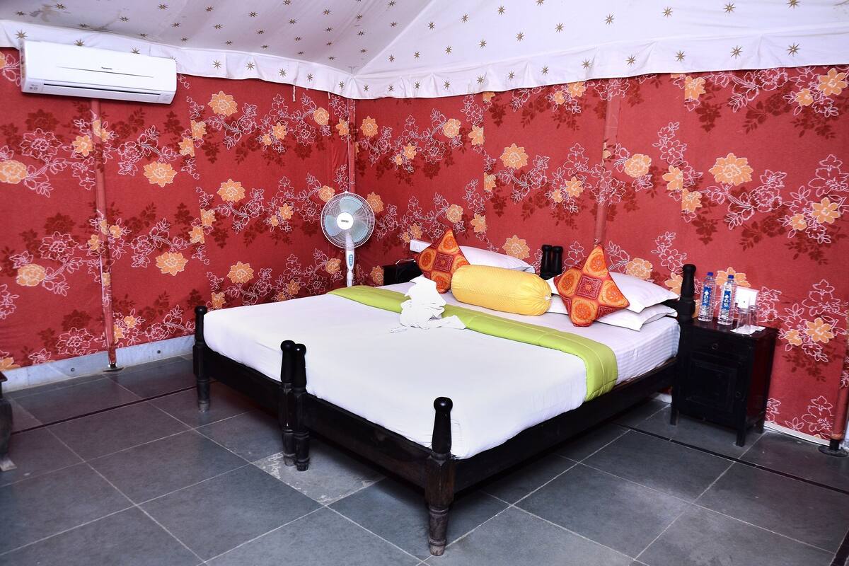 A neatly made bed with a white and green bedspread, a standing fan, and a nightstand with toiletries.