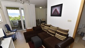 A living room with a brown sofa, blue chairs, a dining table, and a view through a sliding glass door. - Apartment Gloria (Dobrinj)
