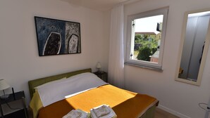 A neatly arranged hotel room with a bed, a bedside table with a lamp, a window with a view of the outside, and a painting on the wall. - Apartment Gloria (Dobrinj)