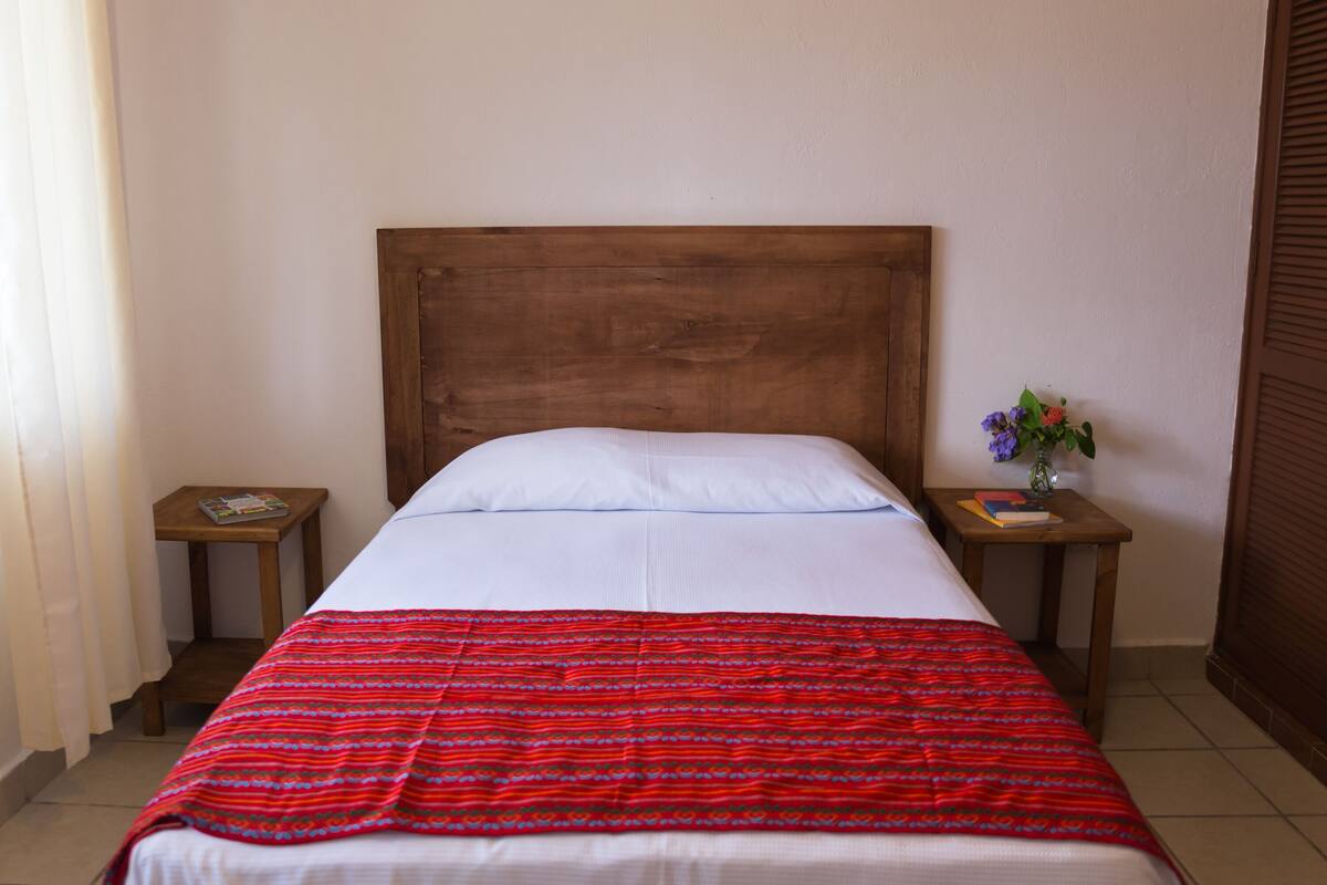 Una cama individual con cabecera de madera, ropa de cama blanca y una manta roja con estampado. Hay dos mesitas de noche de madera, una con un libro y un pequeño jarrón con flores.