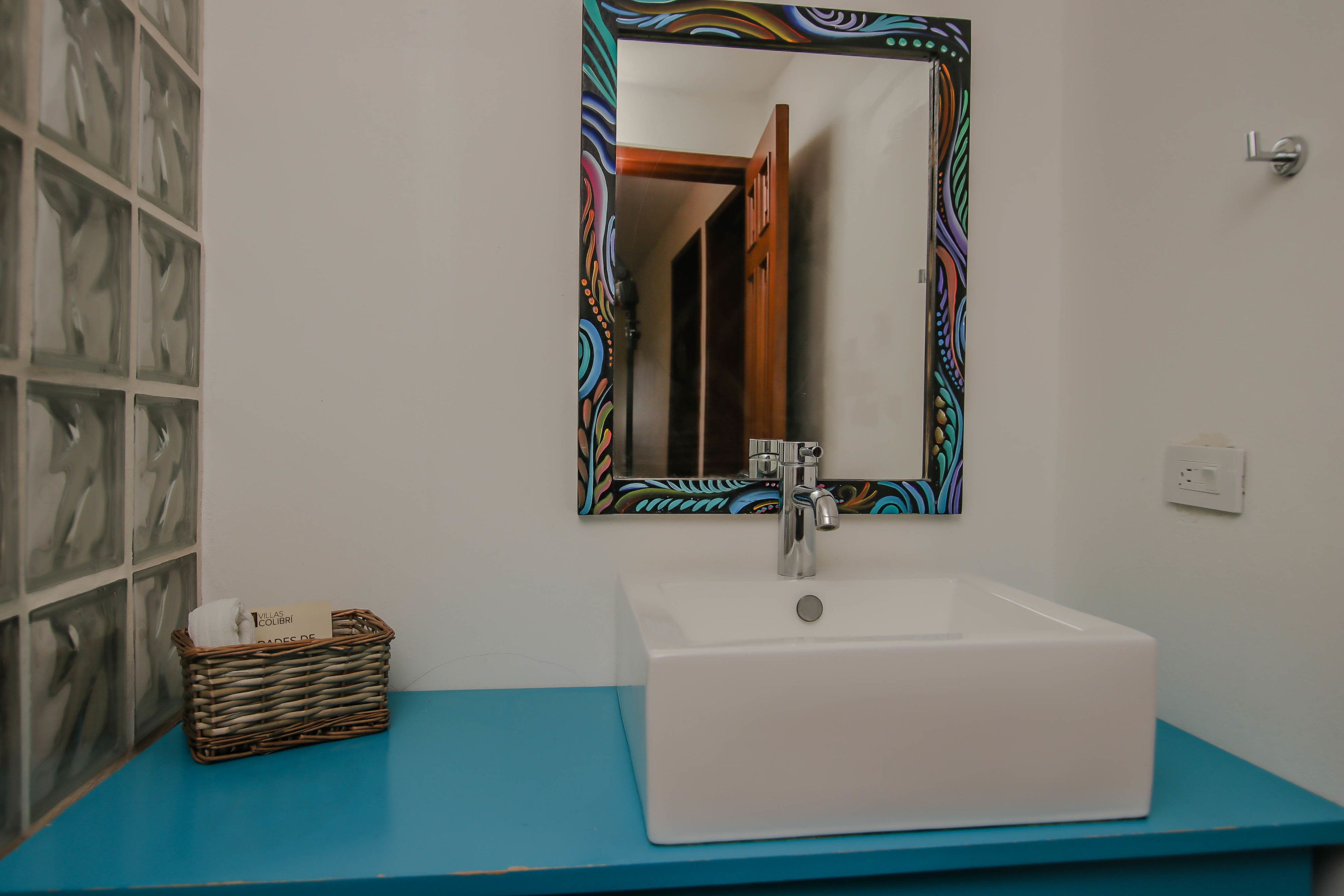 A bathroom with a white sink, a blue countertop, a decorative mirror, and a wicker basket with a tissue.