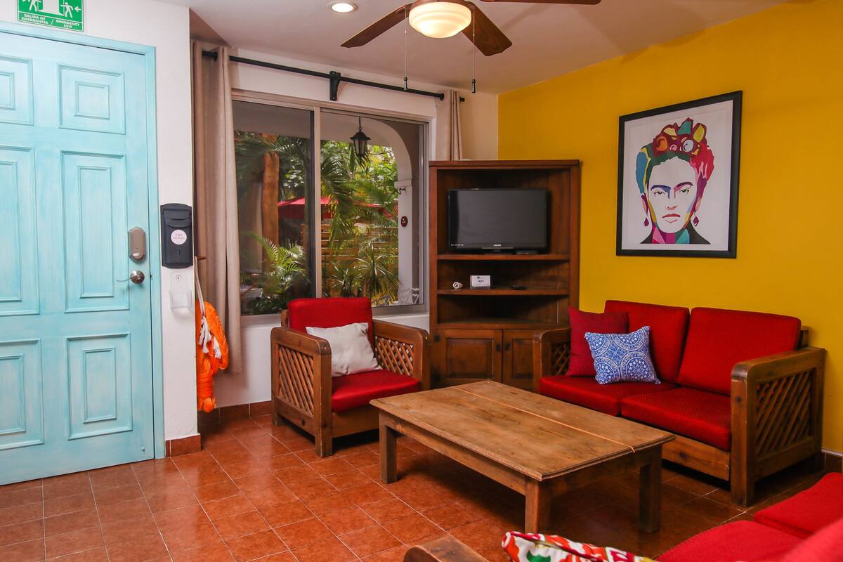 Una habitación con una puerta azul, un televisor sobre un mueble de madera, un sofá rojo, una mesa de centro de madera y un cuadro grande en la pared.