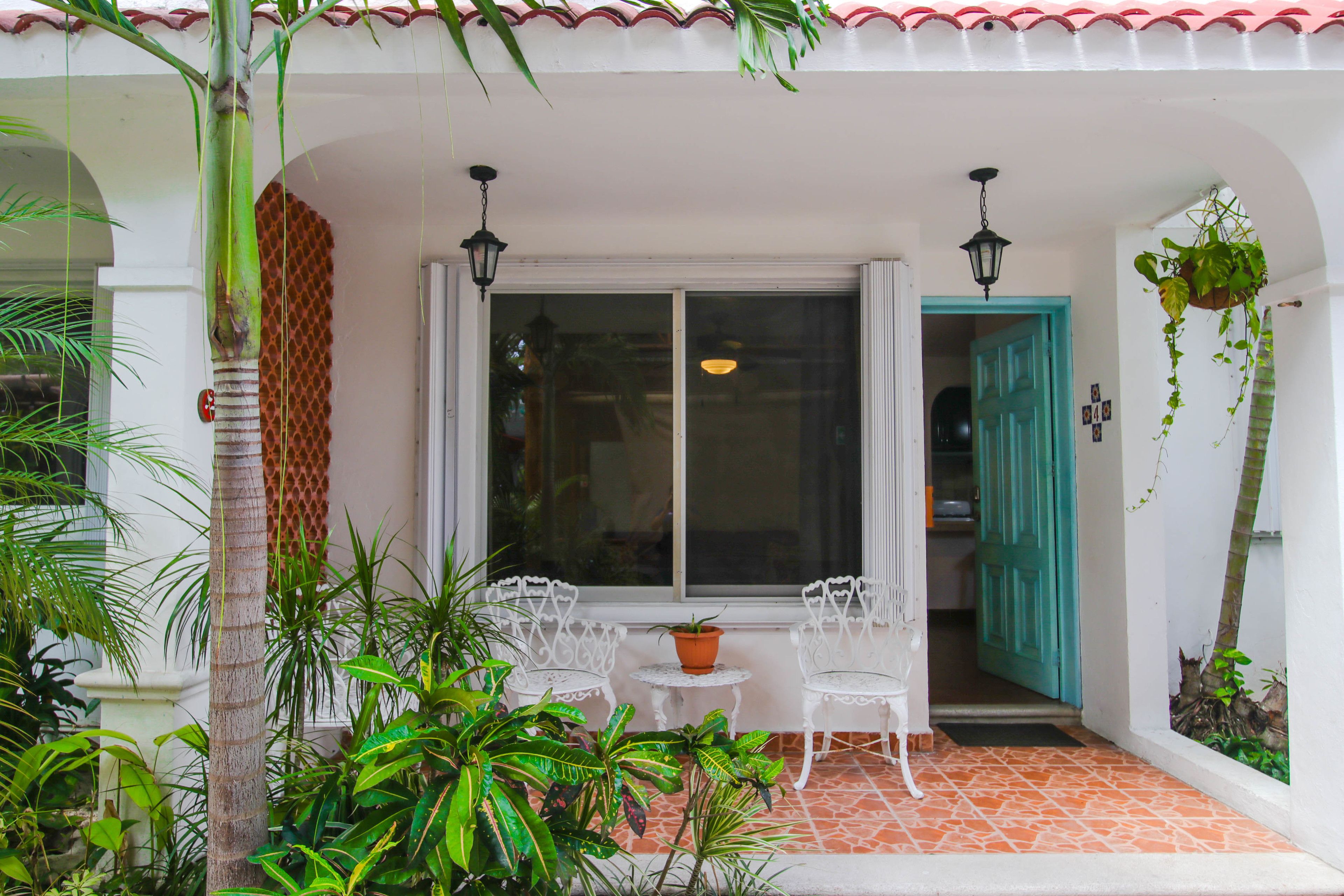 Un espacio de estar al aire libre cubierto, con sillas blancas y una mesa, rodeado de plantas en macetas y una palmera.