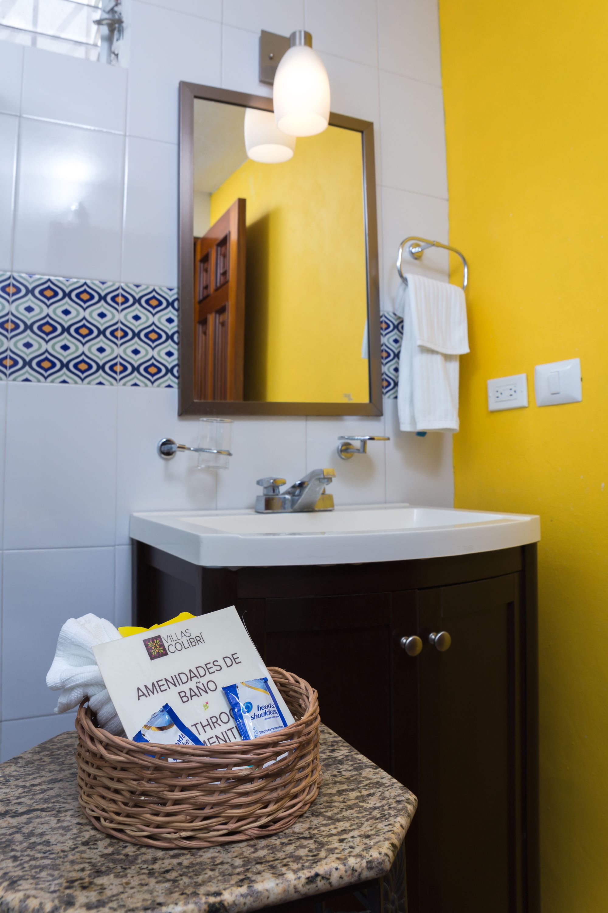 Une salle de bain avec un lavabo blanc, un miroir, un mur jaune et un pare-chocs en carreaux à motifs.