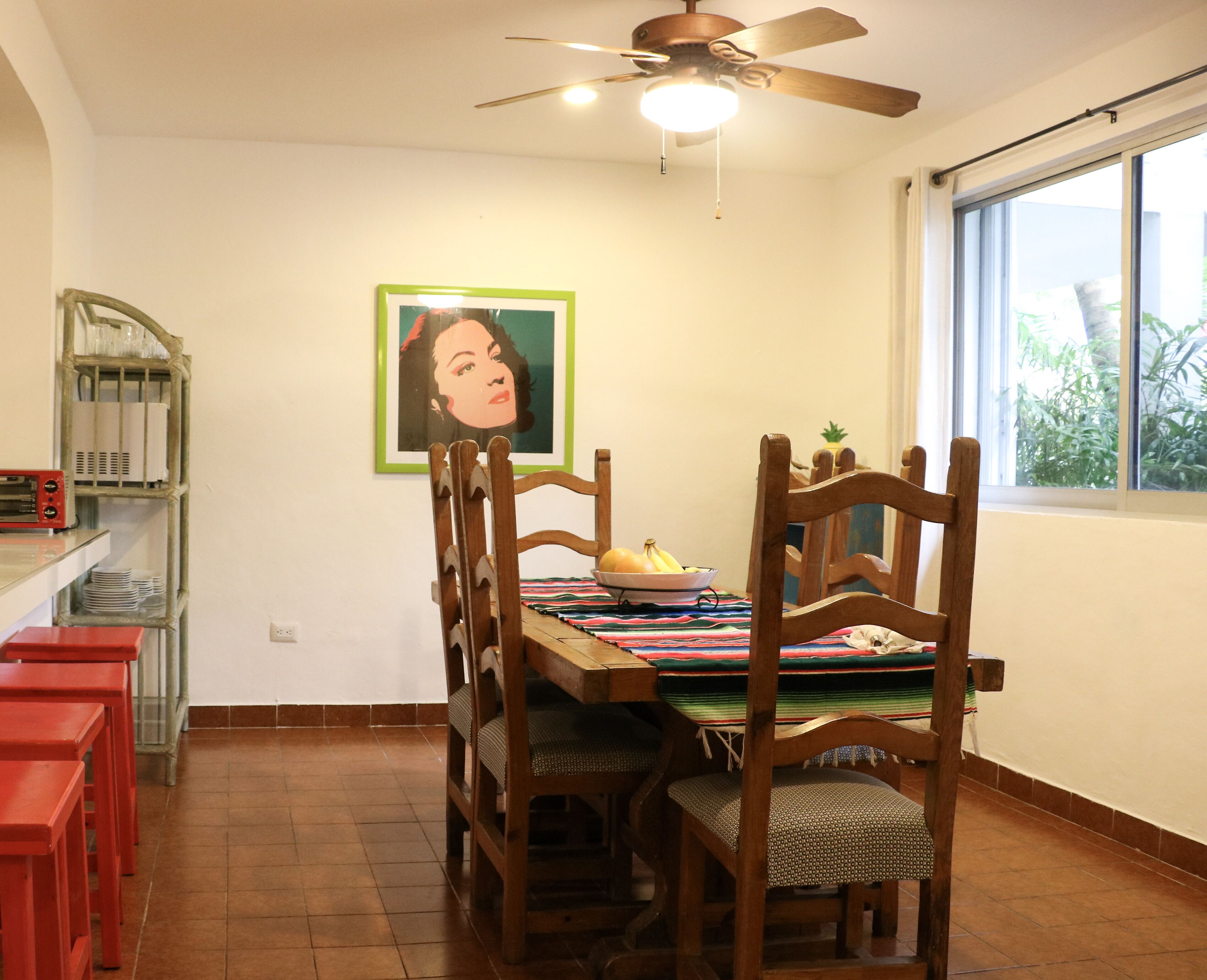 Un coin repas avec une table en bois, des chaises et un ventilateur de plafond. Une fenêtre offre une vue sur la verdure à l’extérieur.