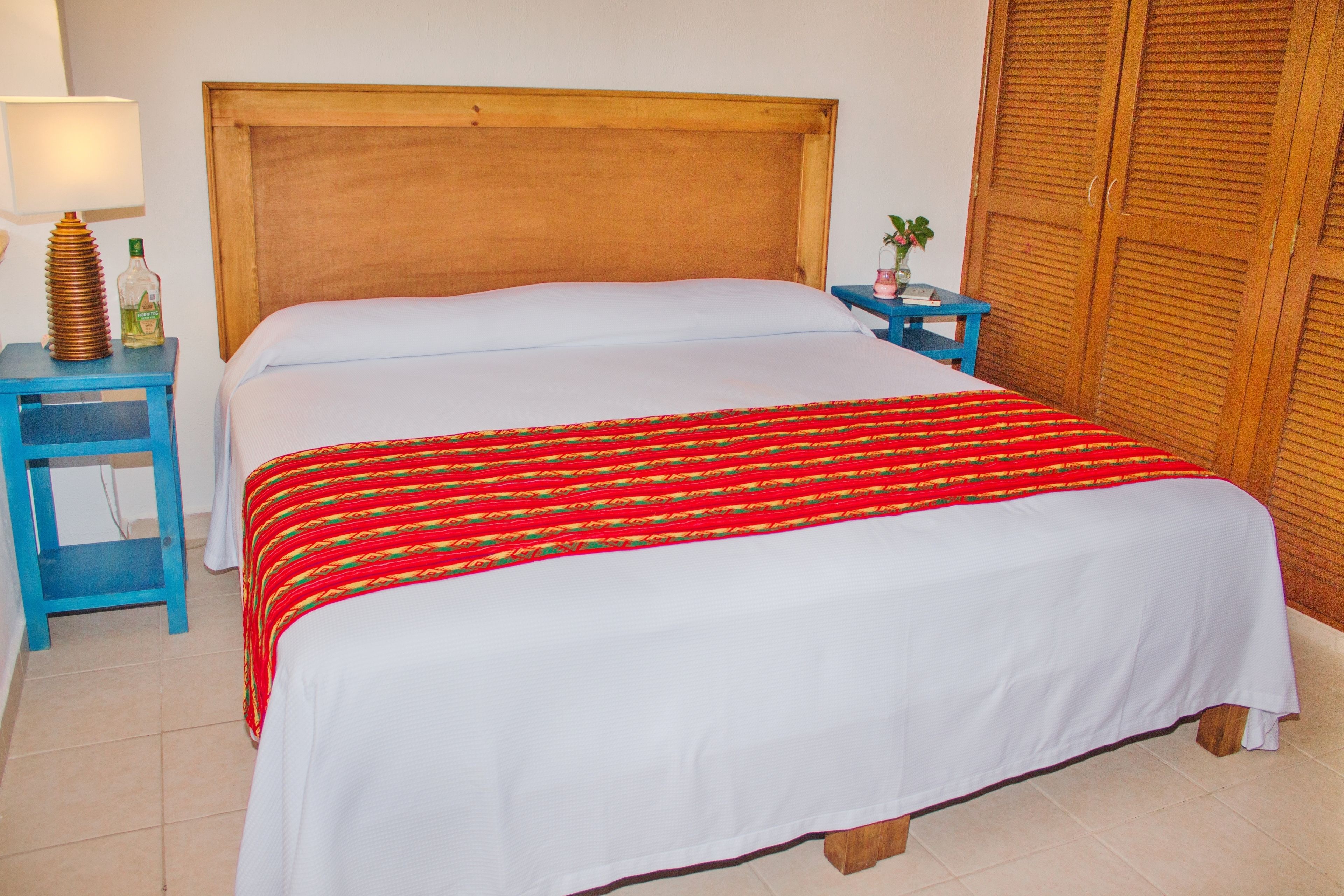 Ein Bett mit einem Holz-Kopfstück, einer rot-gelb gestreiften Decke und einem weißen Überwurf. Neben dem Bett steht ein kleiner Tisch mit einer Lampe und einer Wasserflasche.
