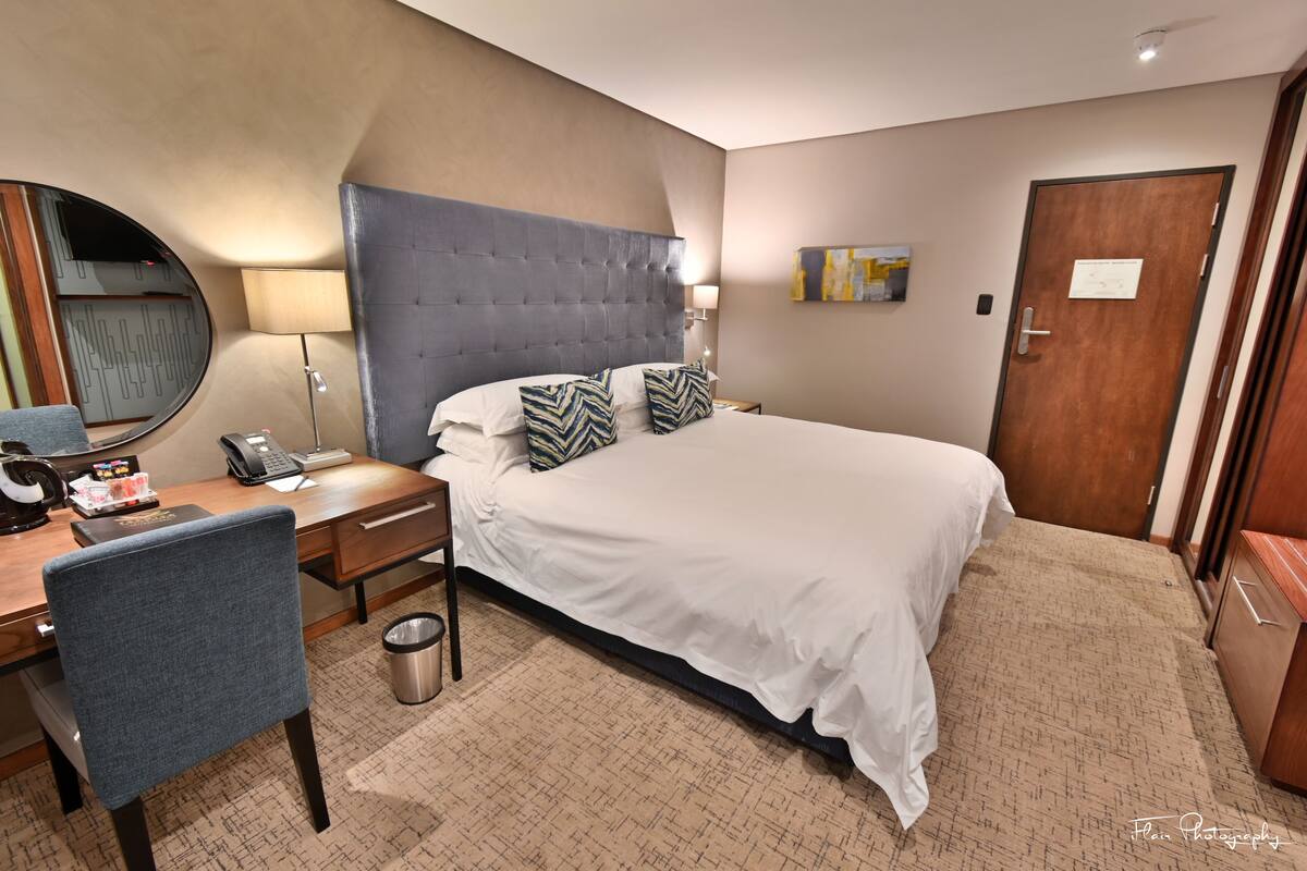 A hotel room with a large bed, a desk with a chair, a telephone, a trash bin, and a door.