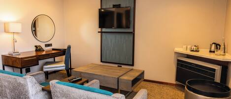 Habitación de hotel con escritorio, silla, sofá, televisor, espejo, cesto de basura y kitchenette.