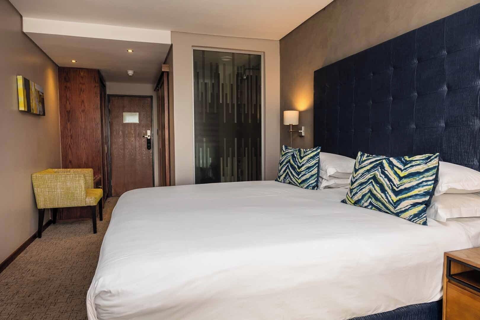 A hotel room with a large bed, a wooden chair, a glass door, and a dark headboard.