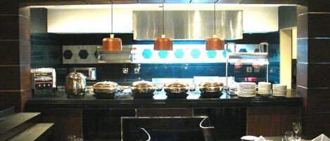 Cocina de restaurante con electrodomésticos de acero inoxidable, una isla central con alimentos y luces colgantes.
