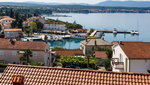 Eine Küstenstadt mit terrakottafarbenen Dächern, einem Yachthafen mit Booten und Blick auf das Meer und ferne Berge.