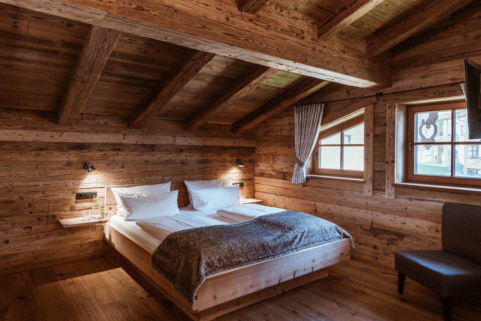 A wooden cabin bedroom with a bed, a chair, and a window with curtains.