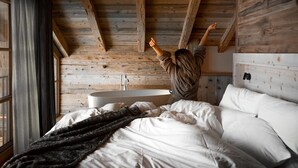 A bedroom with a wooden ceiling, a large bed with white linens, a dark throw blanket, and a headboard with a mounted mirror. - Alpzitt - Chalets (Burgberg)