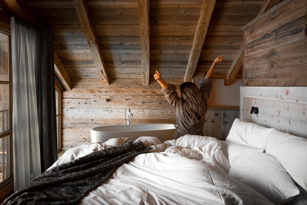 Un dormitorio con techo de madera, una cama grande con ropa blanca, una manta oscura y un cabecero con espejo incorporado.