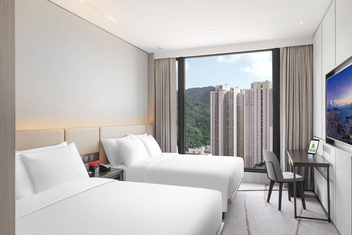 A hotel room with two beds, a desk, and a large window with a city view.