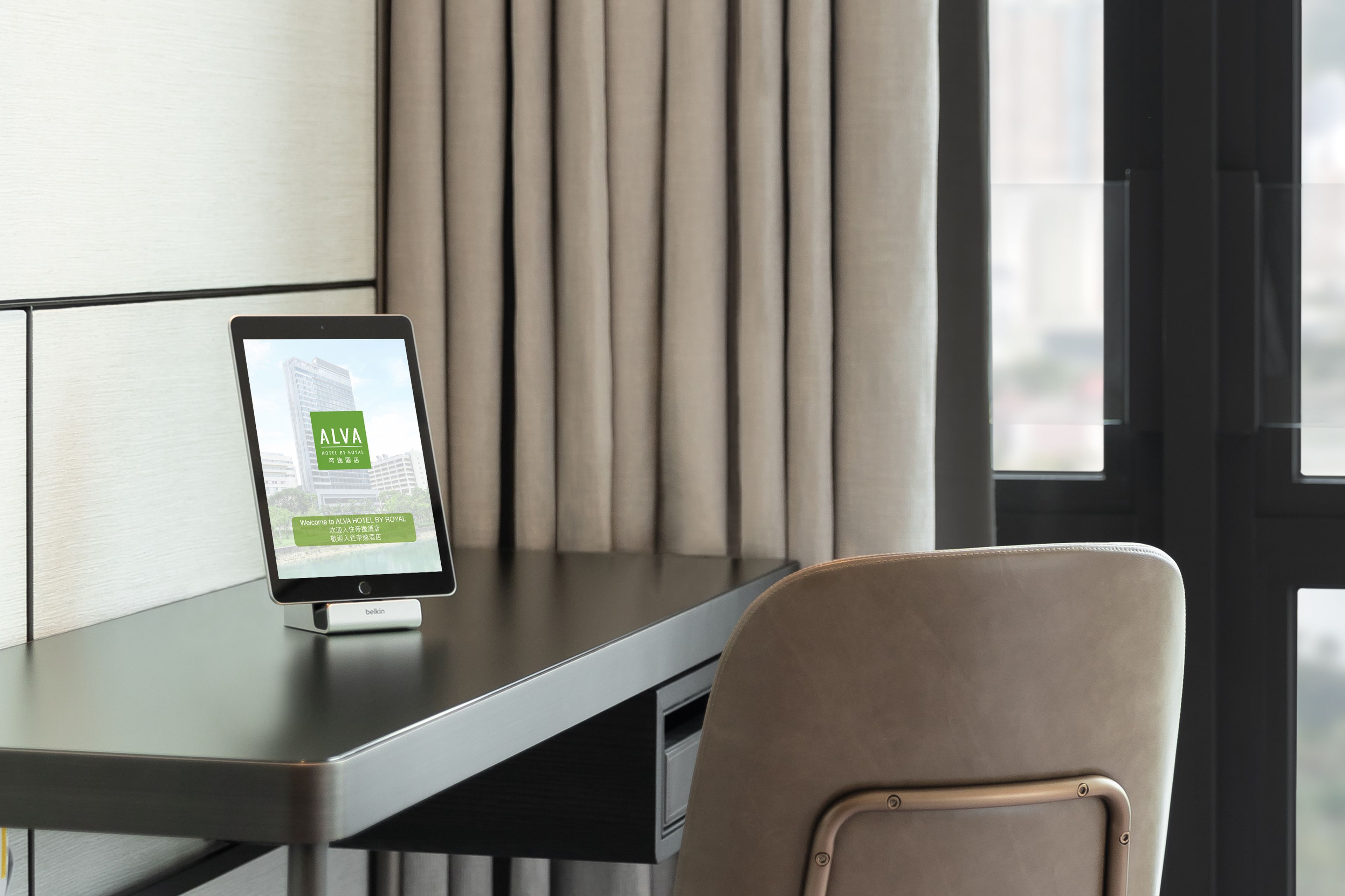 A tablet on a desk displaying a hotel welcome screen, a chair, and a window with curtains.