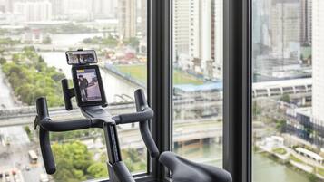 Ein stationäres Fahrrad mit einem Smartphone, das auf einem Halter befestigt ist, steht vor einem Fenster mit Blick auf eine Stadtlandschaft, einschließlich Gebäude und Fluss.