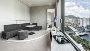 A modern hotel room with a grey sectional sofa, a small round table, and a view of a cityscape and river. - Alva Hotel By Royal (Sha Tin)