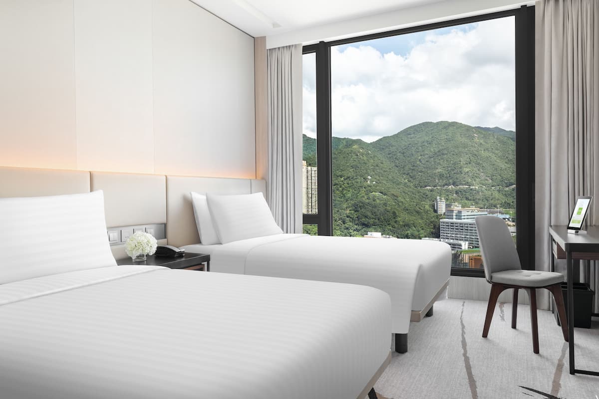 A hotel room with two beds, a desk, and a chair. The room has a large window with a view of a mountainous landscape.