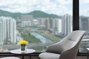 Habitación de hotel moderna con vistas a un río y al paisaje urbano, una butaca tapizada en gris y una mesita con un jarrón de flores amarillas.