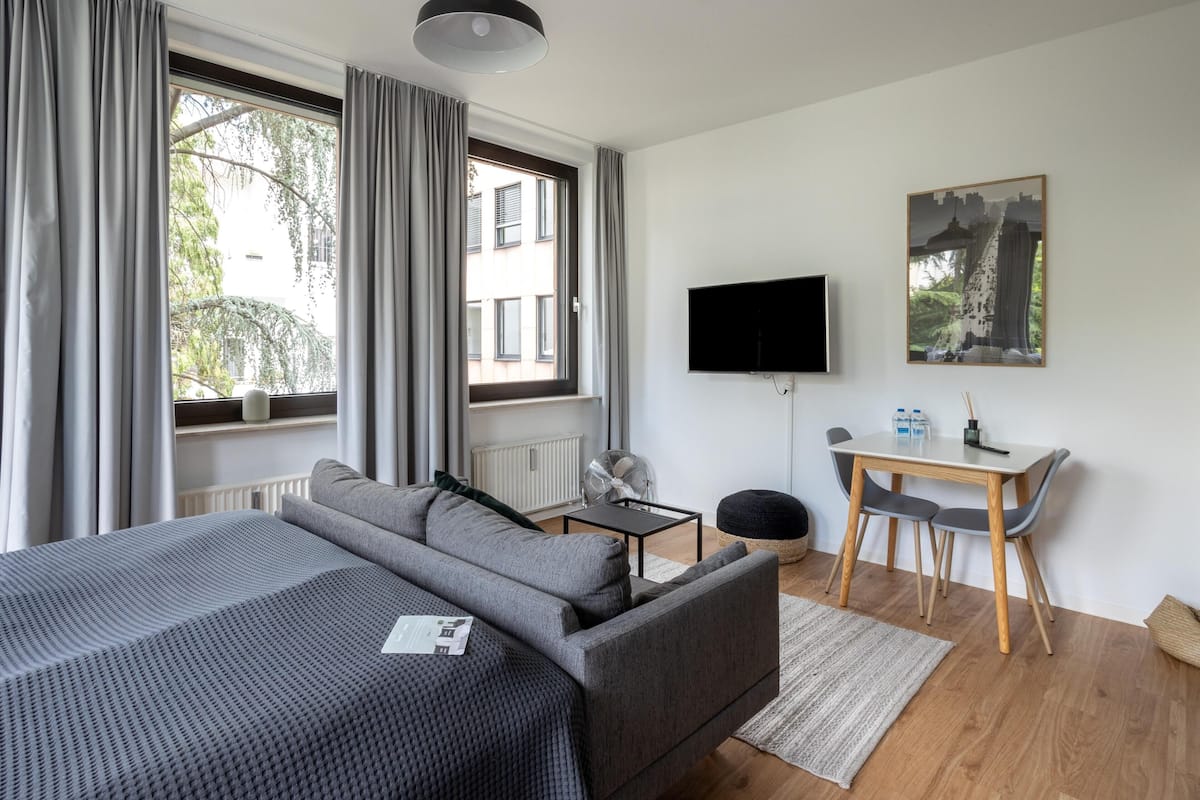 Een moderne kamer met een bank, een aan de muur bevestigde televisie, een kleintje tafeltje met stoelen en een raam met gordijnen.