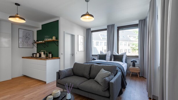 A modern apartment with a grey sofa, a bed, a small kitchen area, and large windows offering a view of buildings.