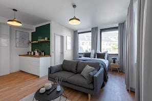 A modern apartment with a grey sofa, a bed, a small kitchen area, and large windows offering a view of buildings. - Limehome Aachen Vereinsstrasse (Aachen)