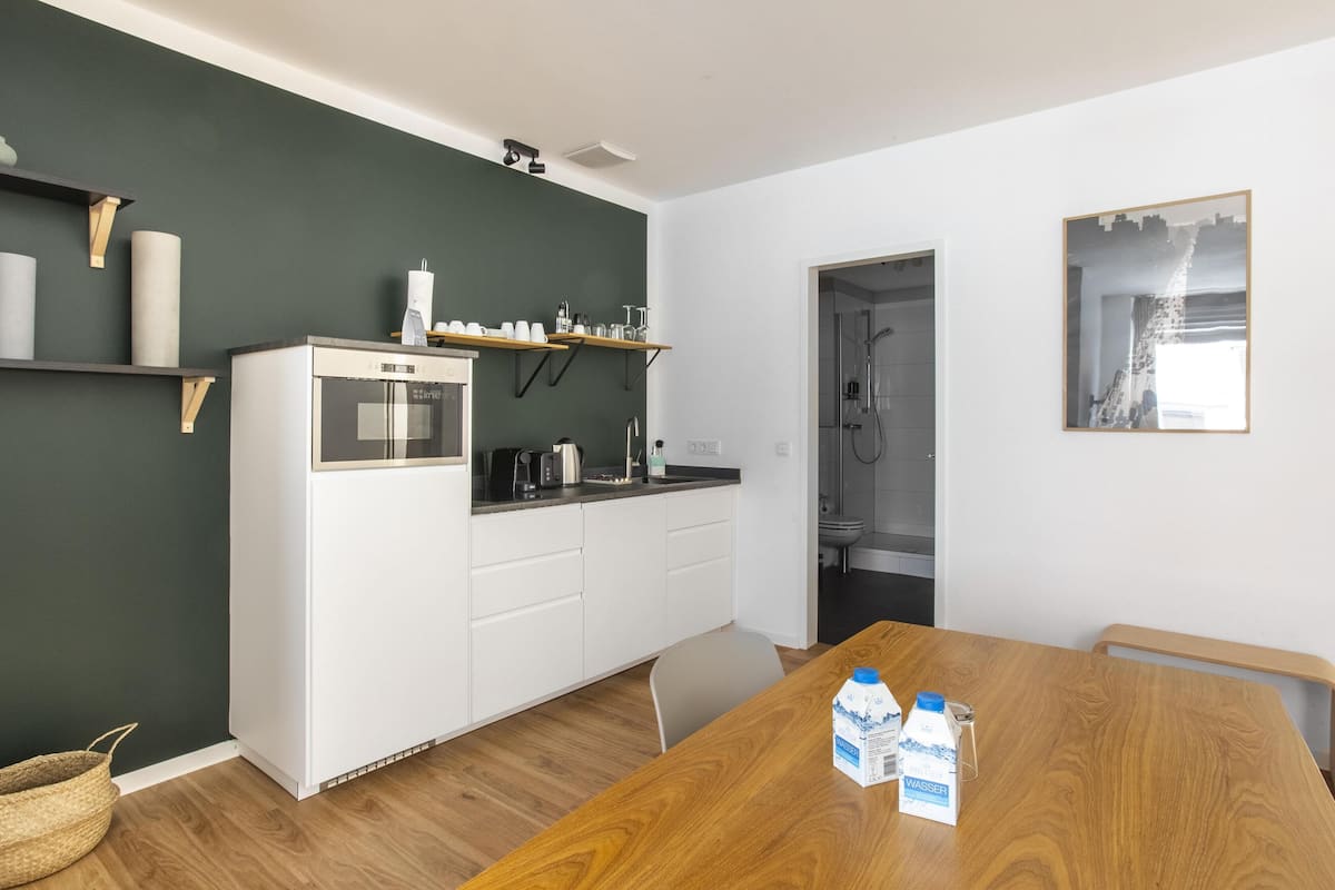 Een compacte keuken met een koelkast, magnetron en koffiezetapparaat, een houten eettafel met melkpakken, en een uitzicht op een stadsgezicht door een raam.