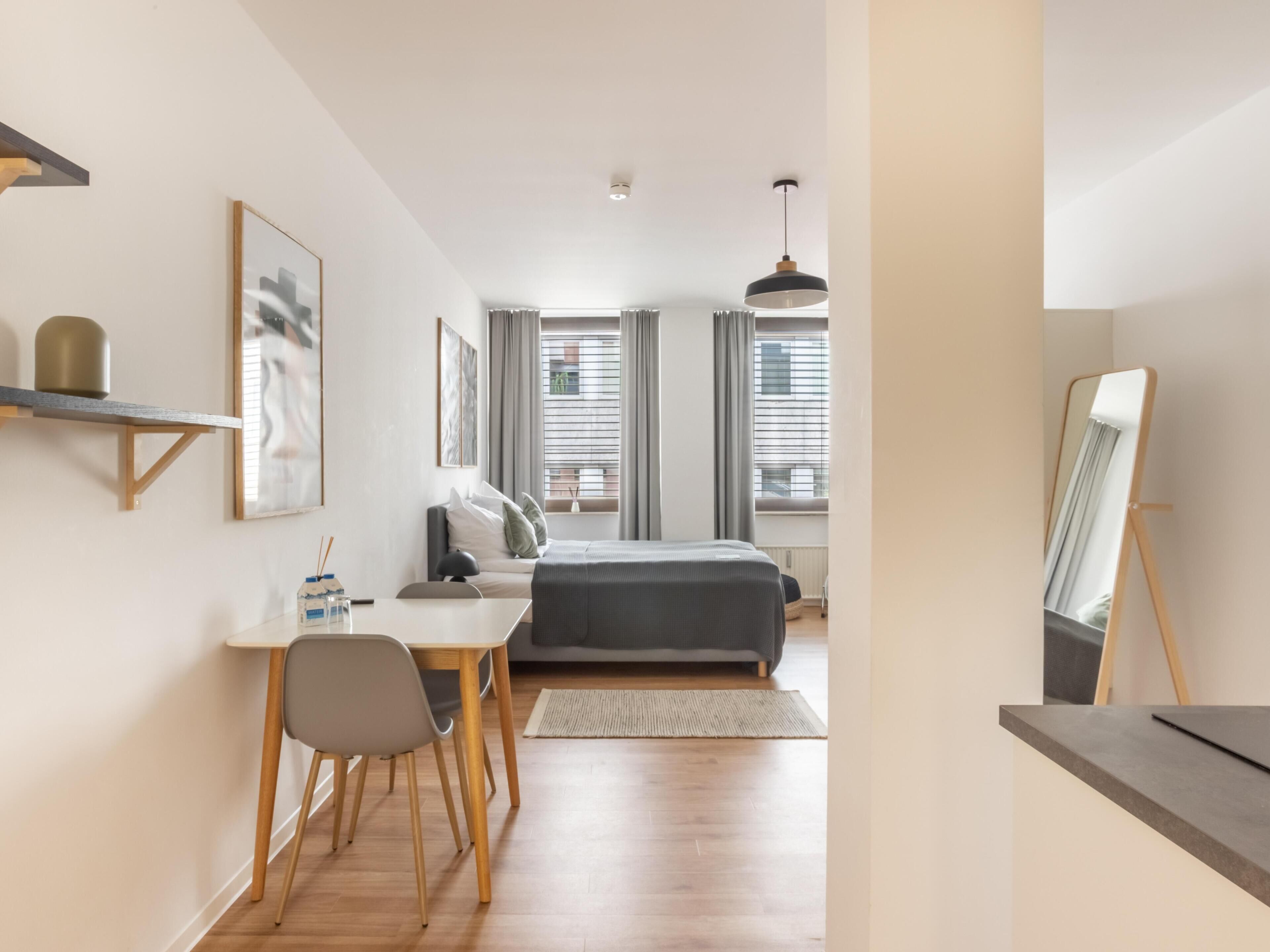 Une chambre moderne comprenant un lit, une table de chevet, une chaise et un bureau avec une autre chaise. Il y a une fenêtre avec des rideaux et une vue sur les bâtiments à l’extérieur.