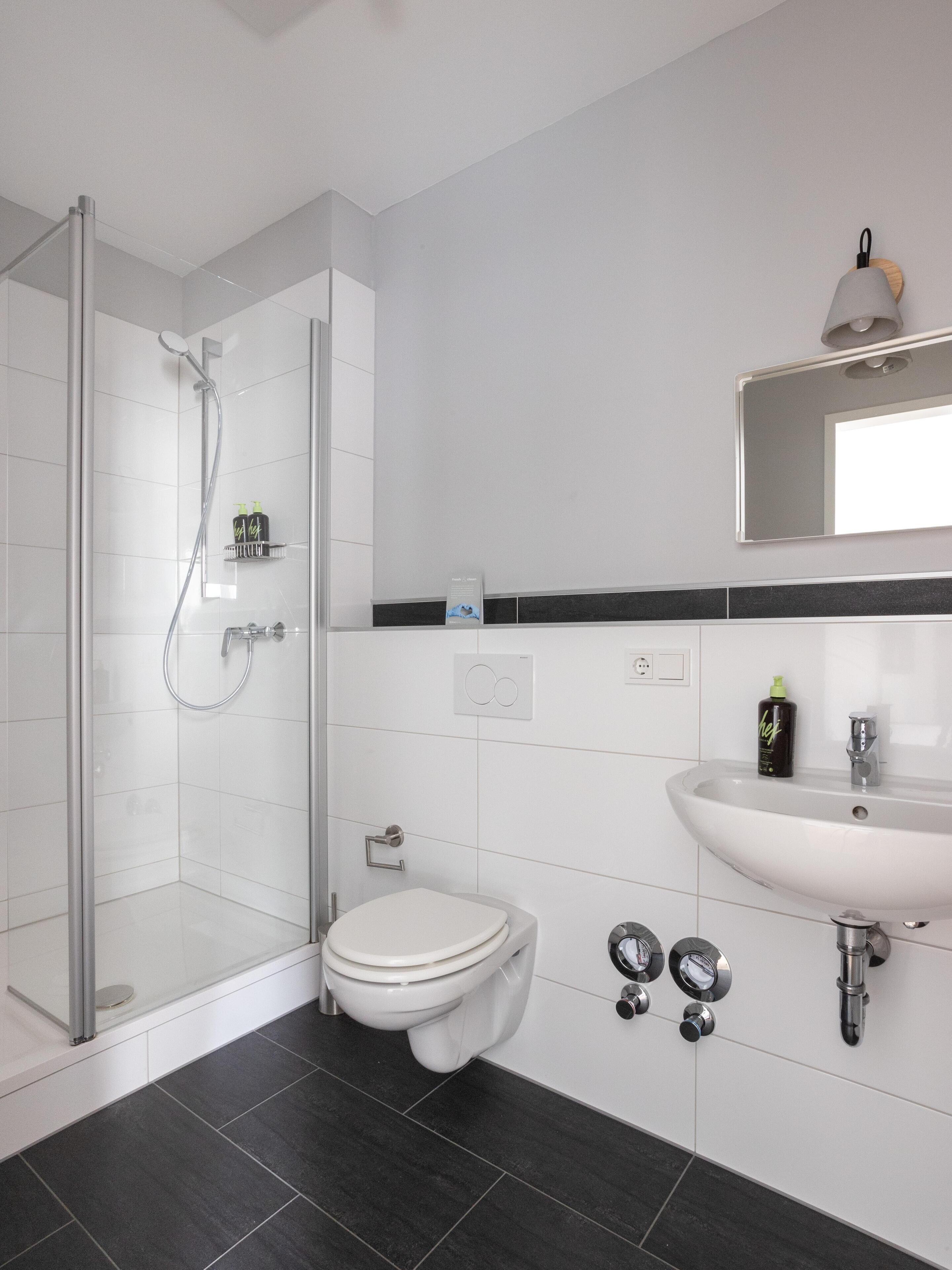 Un baño moderno con cabina de ducha de vidrio, inodoro blanco, lavabo con espejo sobre él y suelo de baldosas negras.