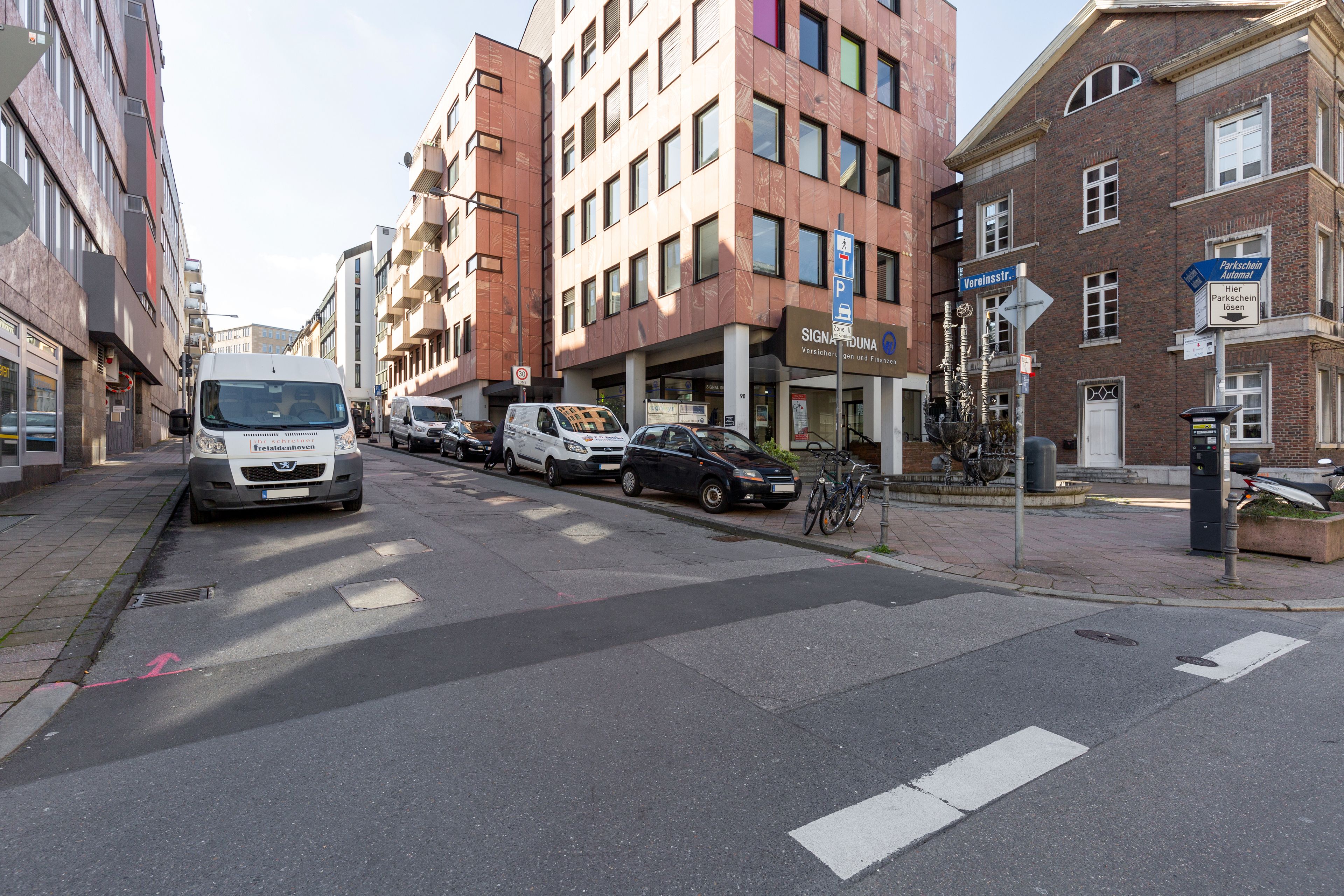 A street with parked cars, a white van, and a building with a sign for 'SIGNATURE HOTEL'.