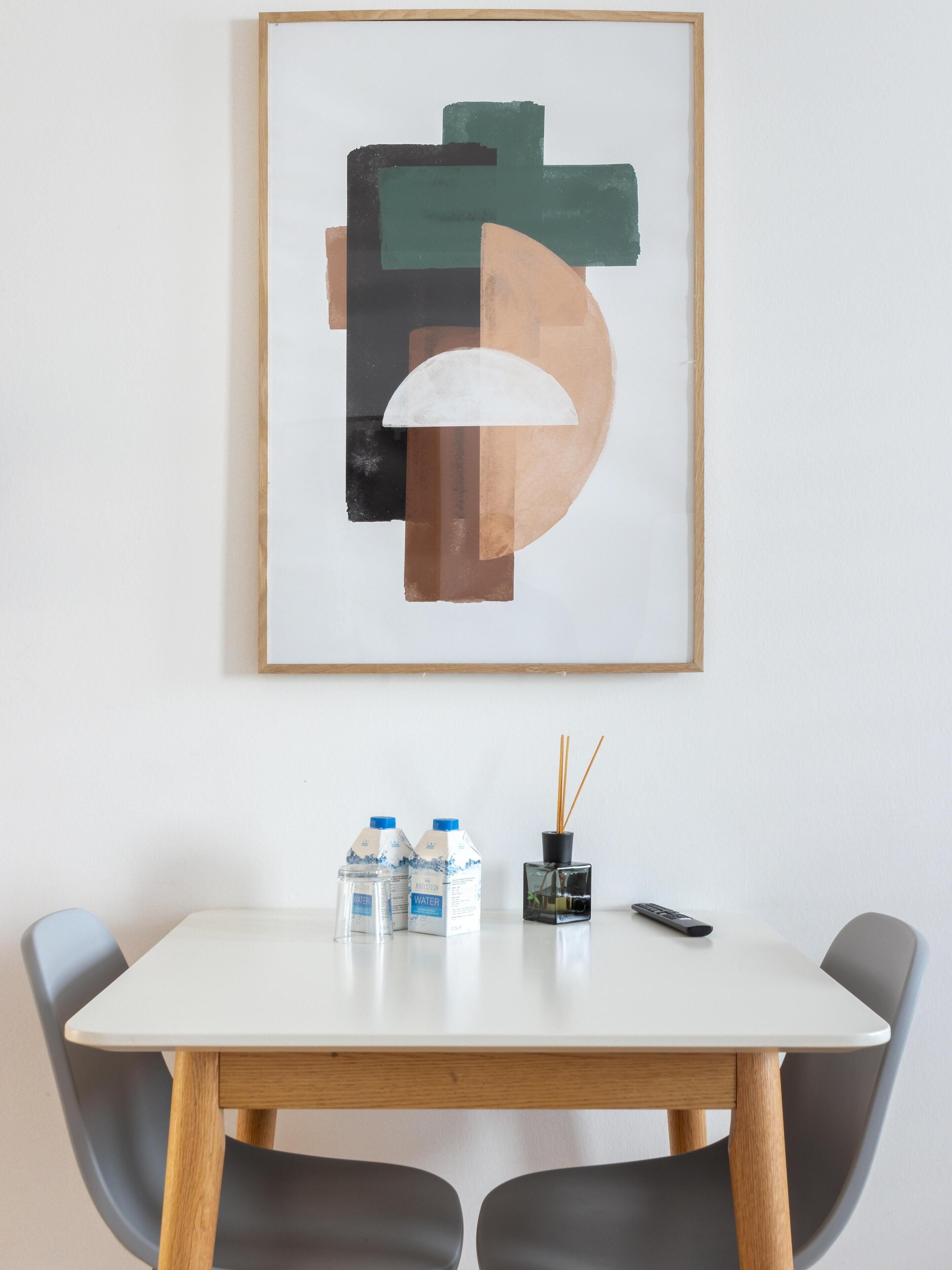 A framed abstract painting on a wall above a white table with two water bottles, a remote control, and a small black vase with sticks.