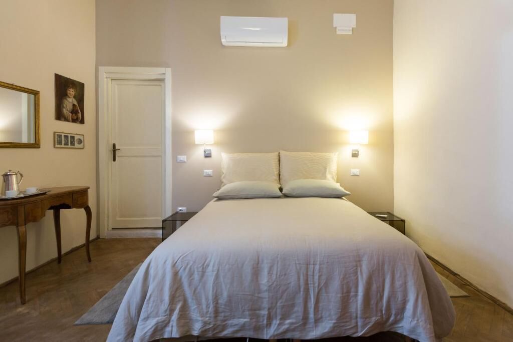 A bedroom with a bed, bedside tables, a desk, a chair, a door, and wall-mounted lights.