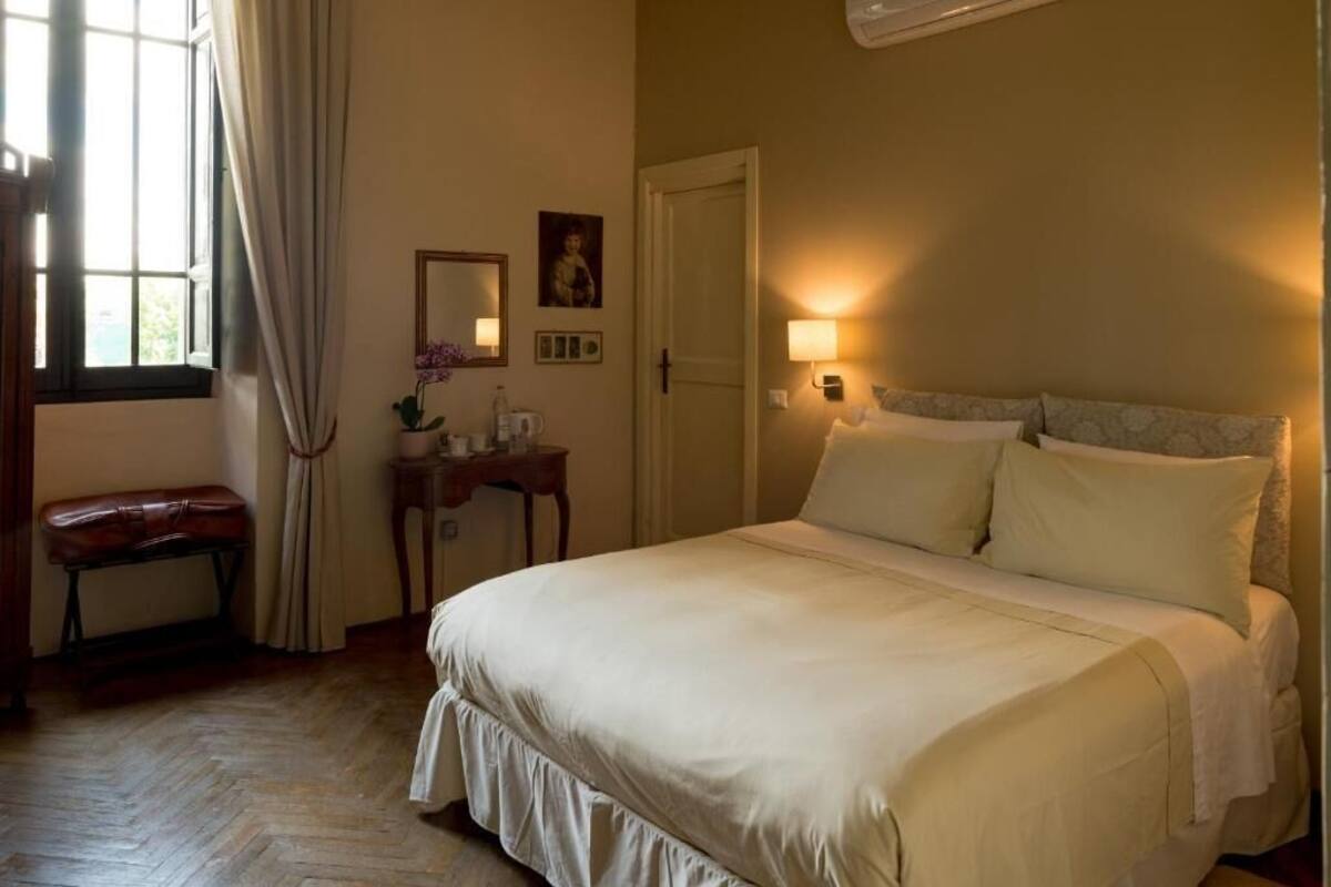 A bedroom with a bed, a bedside table, a lamp, a window with curtains, and a bench.