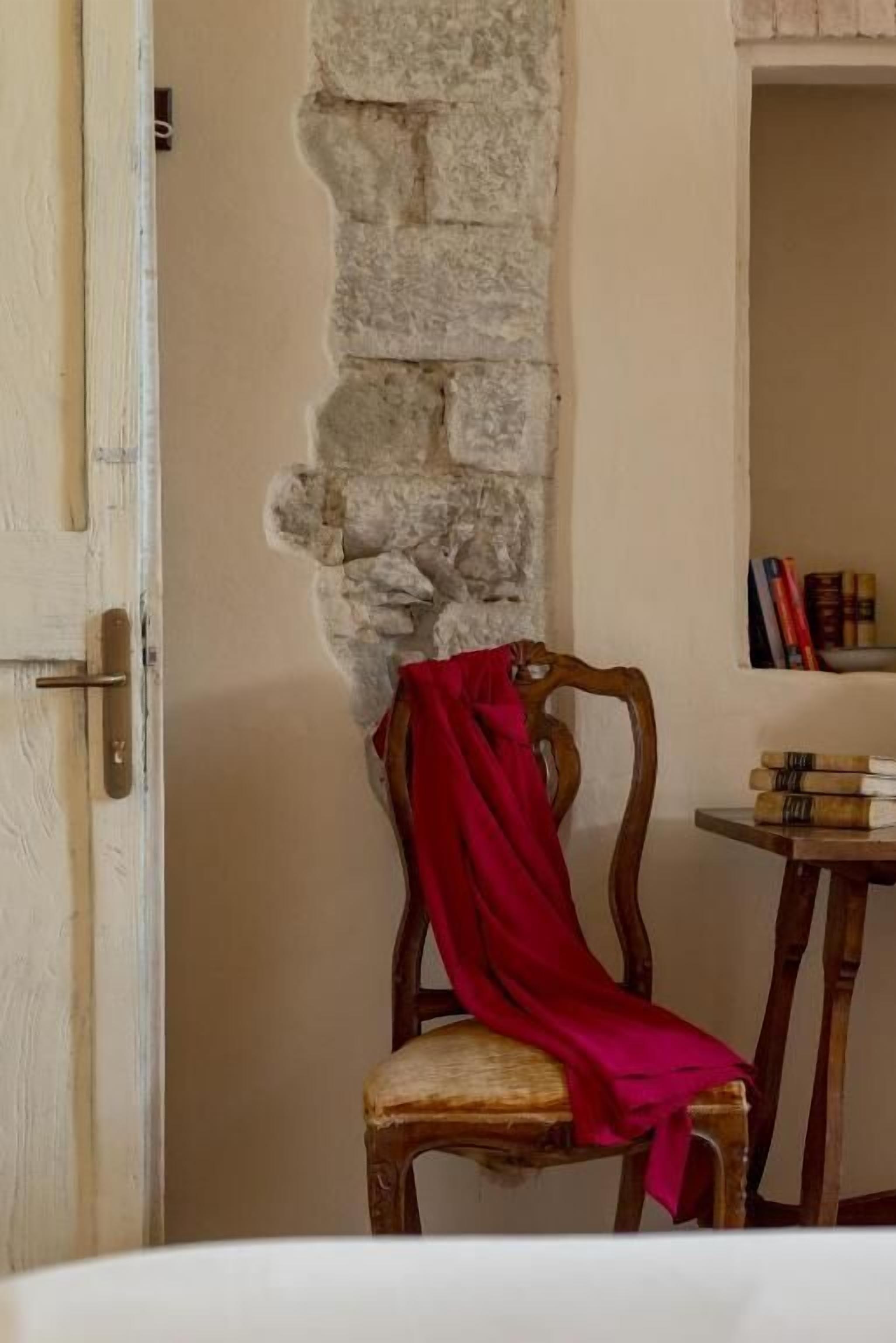 A room with a stone wall, a wooden chair with a red cloth, and a small table with books.