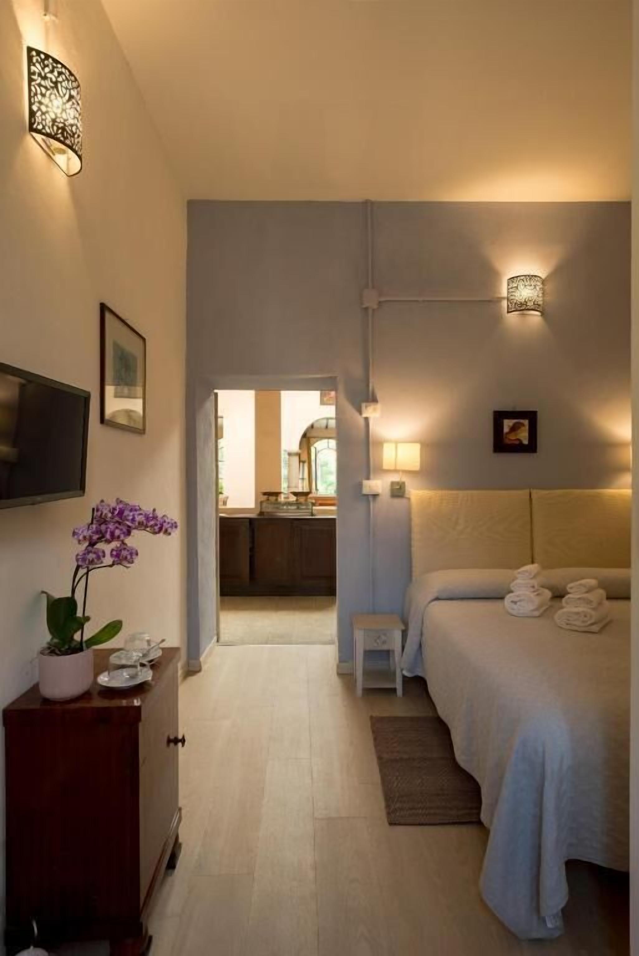 Habitación de hotel con cama, mesita de noche, lámpara y vista a la cocina.