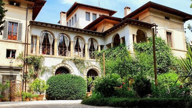 Um edifício de dois andares com janelas em arco, um jardim com diversas plantas e um caminho de cascalho.